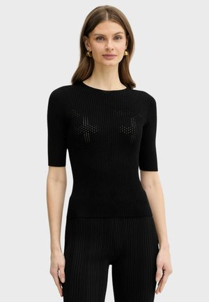 Donna con pelle chiara e capelli castani indossa un top nero a coste aderente a maniche corte e pantaloni a coste abbinati, in piedi davanti a uno sfondo semplice.