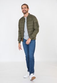 Oliwkowa, pikowana kurtka bomberka z zamkiem błyskawicznym i boczną kieszenią, połączona z szarym t-shirtem i niebieskimi dżinsami, uzupełniona białymi trampkami.