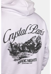 Sweat à capuche blanc avec un graphique noir de montagnes et une cabane, le texte indique "Crystal Paris" et "Nordic Nights Crystal Paris © 2024".