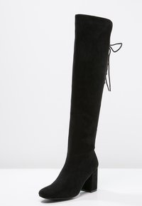 Stivale nero sopra il ginocchio in morbido camoscio, con punta tonda, tacco a blocco e un dettaglio annodato sul retro per una vestibilità regolabile.