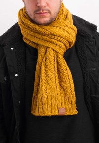 Mustard yellow knitted scarf with a textured cable pattern, frayed ends, and a small leather label. Worn over a black coat.
