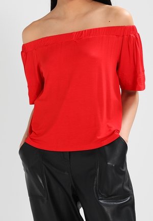 Vrouw met een felrode off-shoulder top met korte mouwen en zwarte leren broek, handen in de zakken, tegen een effen achtergrond.