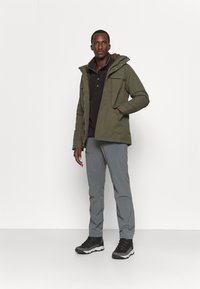 Green insulated jacket with a fur-lined hood, black button-up shirt underneath, gray pants, and dark hiking boots. Standing against a white background.