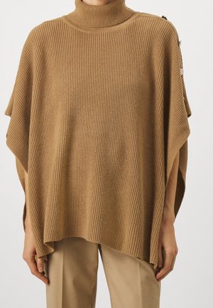 Femme portant un poncho en maille côtelée couleur chameau avec un col roulé et des détails à boutons sur l'épaule, associé à un pantalon beige.