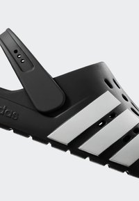 Svart lättviktsklämma med tre vita ränder. Har ett flexibelt band, ventilationshål och en strukturerad sula för grepp.