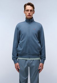 Blauwe rits-sweater met hoge kraag, ribgebreide boorden en zoom. Klein logodat op de borst. Draagt over een lichtgroen shirt en grijze broek.