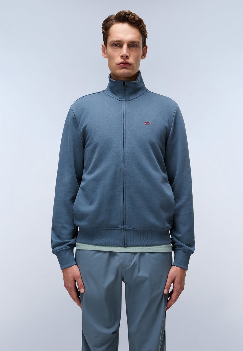 Blauwe rits-sweater met hoge kraag, ribgebreide boorden en zoom. Klein logodat op de borst. Draagt over een lichtgroen shirt en grijze broek.