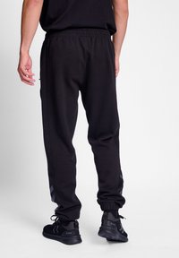 Pantalones de chándal negros con cintura elástica, con rayas laterales y tobillos ajustados. Combinados con zapatillas deportivas negras.