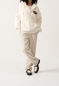 Crèmekleurige hoodie met rits en een logopatch, gecombineerd met beige broek en zwarte schoenen met een geweven ontwerp. Overal een gladde stoftextuur.