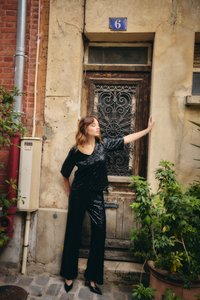 Donna in abito nero con paillettes e tacchi alti, in piedi con un braccio teso, appoggiata a una porta di legno consumata con il numero 6, circondata da piante e muri di mattoni.