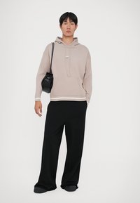 Svetlo bež hoodie s sprednjim žepom, rebrastimi manšetami in kapuco s vrvico, v kombinaciji s ohlapnimi črnimi hlačami in majhno črno torbico.