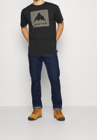 Černé bavlněné tričko s šedým čtvercem, na kterém je grafika hory a text "BURTON". Nosené s tmavě modrými džíny a žluto-hnědými botami.