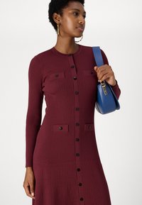 Burgundi bordás ruha hosszú ujjakkal, gombos elülső résszel, két mellzsebbel és fekete gombokkal. Kék táskával párosítva.