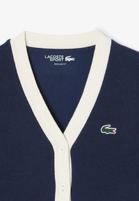 Cardigan navy con scollatura a V a costine color crema e chiusura con bottoni. Presenta il logo coccodrillo Lacoste verde e rosso sul lato sinistro. Vestibilità regolare.