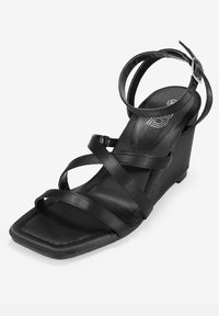 Sandalia cuña negra con diseño de punta cuadrada, adornada con tiras entrelazadas de material de satén suave. Cuenta con una correa en el tobillo con un hebilla.