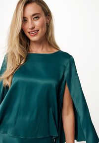 Top in raso teal con scollatura quadrata e maniche fluide. Presenta una texture liscia e dettagli a taglio sui lati.