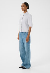 Heled hall kardigan lühikeste varrukatega, kõrge kaelusega ja nupuga kinnitusega, koos vabade siniste teksade ja terava musta jalatsiga.