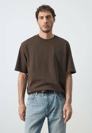 Mann mit kurzen lockigen Haaren, der ein braunes kurzärmeliges Hemd trägt, das in eine hellblaue Jeans mit schwarzem Gürtel gesteckt ist, steht vor einem einfarbigen Hintergrund.