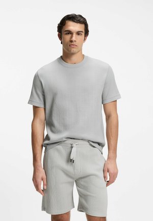 Junger Mann trägt ein hellgraues, strukturiertes, kurzärmeliges Strickshirt und passende Shorts mit Kordelzug, steht vor weißem Hintergrund.