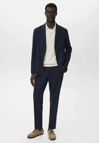Marineblazer mit zwei vorderen Taschen, cremefarbenem Poloshirt, marineblauen Hosen und beigen Schnürschuhen. Glattes Gewebe mit taillierter Passform.