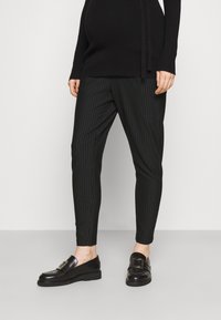 Person in schwarzen, gestreiften, tapered Hosen, schwarzen Loafers mit Metall-Schnallen und einem schwarzen, langärmligen Oberteil, steht vor einem einfarbigen Hintergrund.