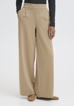 Woman wearing beige wide-leg trousers with pockets, paired with brown flat shoes and a long-sleeve black top, standing on plain background.