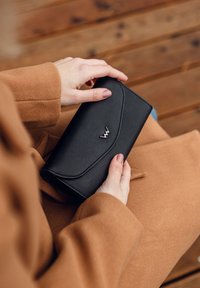 Černá texturovaná peněženka držená v rukou, s klopou ve tvaru vlny a stříbrným logo akcentem. Umístěná na hnědém kabátu a dřevěném pozadí.