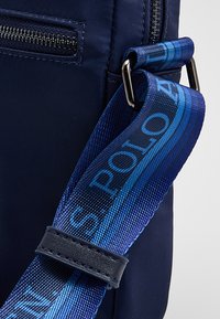 Laivastonsininen nailonlaukku, jossa on vetoketjullinen tasku ja harmaa sinisen sävyinen kudottu hihna, jossa toistuu "Polo"-kuvio.