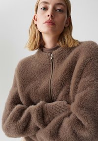 Jonge vrouw met blond haar die een bruin, harig vest met rits en een bijpassende hoog gesloten top draagt tegen een effen lichte achtergrond.