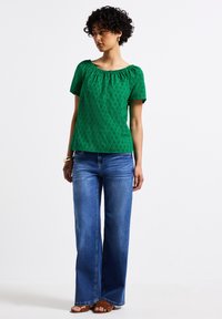 Top verde a maniche corte con motivo testurizzato, scollatura rotonda, abbinato a jeans blu a gamba larga e sandali marroni. Outfit semplice e casual.