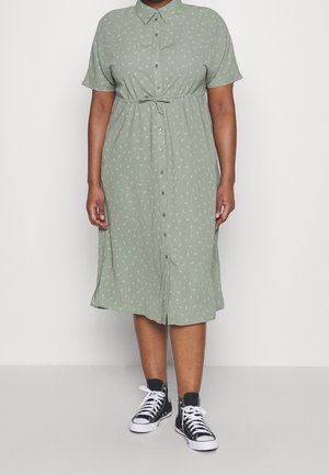 Personne portant une robe verte claire à manches courtes, boutonnée sur le devant, avec une ceinture à la taille, et des baskets montantes noires aux lacets blancs.