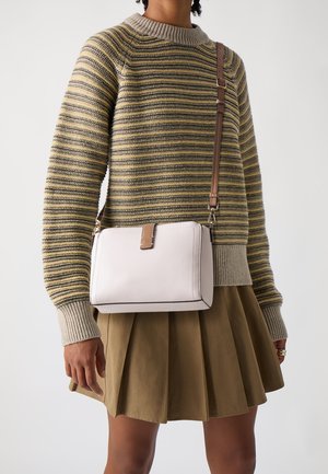 Persona con un suéter de punto a rayas y una falda plisada verde oliva, llevando una bolsa blanca cruzada con correa y cierre marrón.