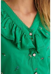 Blusa in cotone verde con scollo arricciato e ricami floreali. Chiusura con bottoni rotondi, rifinita con pizzo.