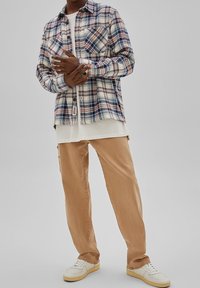 Uomo in camicia a quadri blu, rossa e bianca, indossata sopra una t-shirt bianca, pantaloni beige e sneakers bianche, in piedi davanti a uno sfondo grigio uniforme.