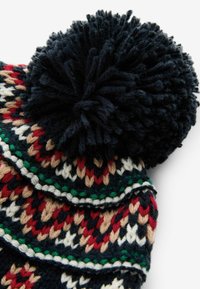 Bonnet tricoté dans des motifs bleu marine, rouge, crème et vert avec un grand pompon sombre sur le dessus. Présente un design géométrique multicolore.