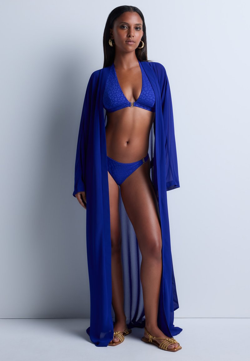 Ensemble bikini bleu avec un motif festonné, des accents dorés et un paréo bleu transparent. Le mannequin se tient sur des sandales à lanières sur un fond neutre.