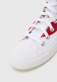 Άσπρο αθλητικό παπούτσι από καμβά με κόκκινη λεπτομέρε�ια, επίπεδη ελαστική σόλα και ασημί οπές. Διαθέτει στρογγυλή μύτη και κλασικό δέσιμο με κορδόνια.