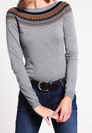 Frau trägt einen eng anliegenden grauen Pullover mit einem mehrfarbigen, gemusterten Einsatz, dunkle Jeans und einen schwarzen Gürtel mit einer silbernen Schnalle.