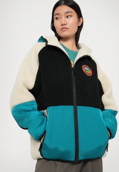 Jachetă cu fermoar căptușită cu fleece, având partea superioară neagră și crem și secțiunea inferioară teal. Include un logo cusut pe față și manșete elastice.
