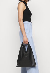 Czarna skórzana torebka o trójkątnym kształcie, z marszczonym uchwytem i srebrnym akcentem. Noszona przez osobę w jasnoniebieskich spodniach.