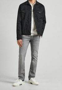 Uomo che indossa una giacca di jeans nera sopra un polo bianco, jeans grigi chiari e scarpe da ginnastica bianche, in piedi davanti a uno sfondo semplice.