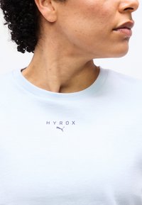 Άτομο με σγουρά μαλλιά που φορά ανοιχτό μπλε πουκάμισο με τυπωμένο το λογότυπο "HYROX" και το λογότυπο Puma στο στήθος, στραμμένο προφίλ.