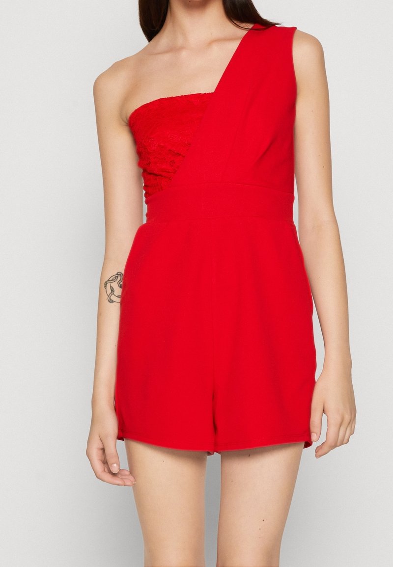 Femme portant un combishort rouge vif à une épaule, avec un tissu texturé sur le corsage et le short, montrant un tatouage sur son avant-bras gauche.