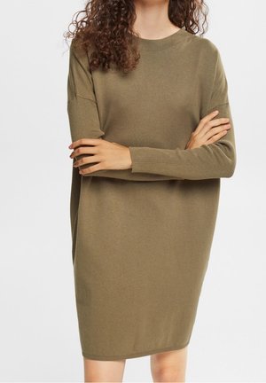 Femme aux cheveux bouclés portant une robe-pull ample en maille vert olive à manches longues, debout les bras croisés.