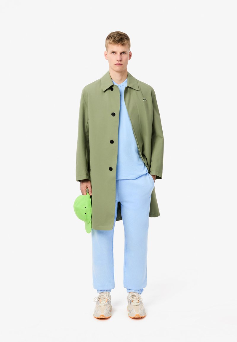 Zielony długi płaszcz z czarnymi guzikami, jasnoniebieski sweatshirt, dopasowane joggery i beżowe sneakersy. Trzyma jasnozieloną czapkę.