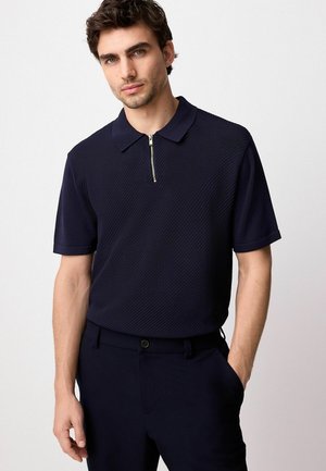 Polo de marina con mangas cortas, tejido texturizado, cuello y detalle de cremallera dorada. Combinado con pantalones navy a juego.