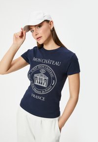 Marineblå bomulds-T-shirt med hvid grafisk tryk, der viser en bygning, tekst og dekorative detaljer. Modellen bærer en hvid kasket.