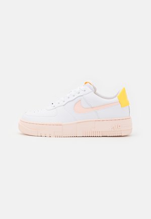 Hvid sneaker med lyst pink sål og Nike swoosh, med en gul hælflap, vist fra siden på en ensfarvet baggrund.