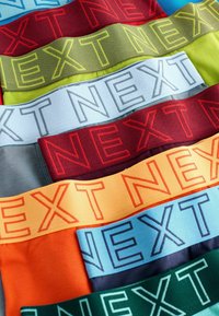 Des bandes de tissu multicolores portant le mot "NEXT" dans différentes polices. Les couleurs incluent le rouge, le vert, le bleu, l'orange, le jaune et le gris.