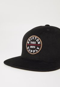 Crni kord kapu s ravnim vizirima, na kojem se nalazi okrugli prošiv s bijelim i crvenim natpisima "Brixton Supply" i "Trade Mark."
